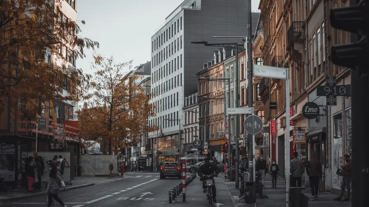 Ulica w Luksemburgu, jesienne drzewa, nowoczesne i zabytkowe budynki. Ludzie idący chodnikiem, rowerzysta, samochody. Praca w Luksemburgu to marzenie wielu.