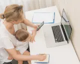 Kobieta z dzieckiem na kolanach sprawdza na telefonie, zasiłek macierzyński ile wynosi, obok laptop i notatnik.