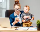 Kto płaci macierzyński? Kobieta w biurze z dzieckiem na rękach, pracująca przy laptopie i telefonie.