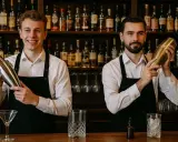 Dwóch młodych mężczyzn na kursie na barmana, każdy z shakerem w dłoni, na tle półek z alkoholem.