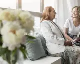 Uśmiechnięta młoda kobieta z książką rozmawia z seniorką przy oknie. Szukam pracy jako opiekunka prywatnie, oferując troskę i towarzystwo.