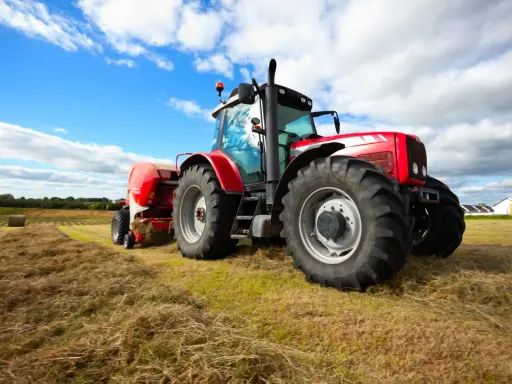 Czerwony traktor z przyczepą prasuje siano na polu. W tle błękitne niebo z chmurami. Traktorzysta w pracy.