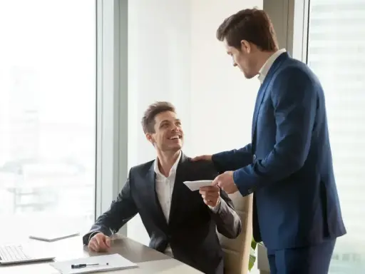 Szef wręcza pracownikowi kopertę z premią uznaniową. Uśmiechnięty mężczyzna siedzi przy biurku, otrzymując zasłużone docenienie.
