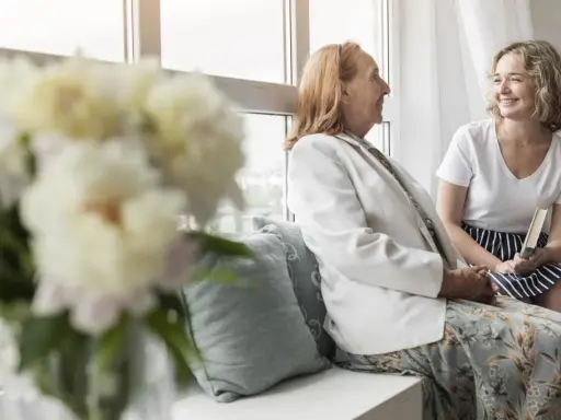Uśmiechnięta młoda kobieta z książką rozmawia z seniorką przy oknie. Szukam pracy jako opiekunka prywatnie, oferując troskę i towarzystwo.