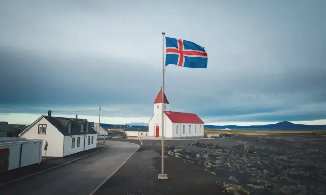 Islandia praca: biały kościół z czerwoną wieżą i flagą Islandii na tle pochmurnego nieba.