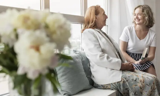Uśmiechnięta młoda kobieta z książką rozmawia z seniorką przy oknie. Szukam pracy jako opiekunka prywatnie, oferując troskę i towarzystwo.