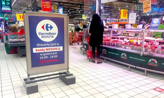Praca w Carrefour: wykładanie towaru na ladę mięsną. W tle widać klientów i tablicę informującą o niedzielnych godzinach otwarcia.