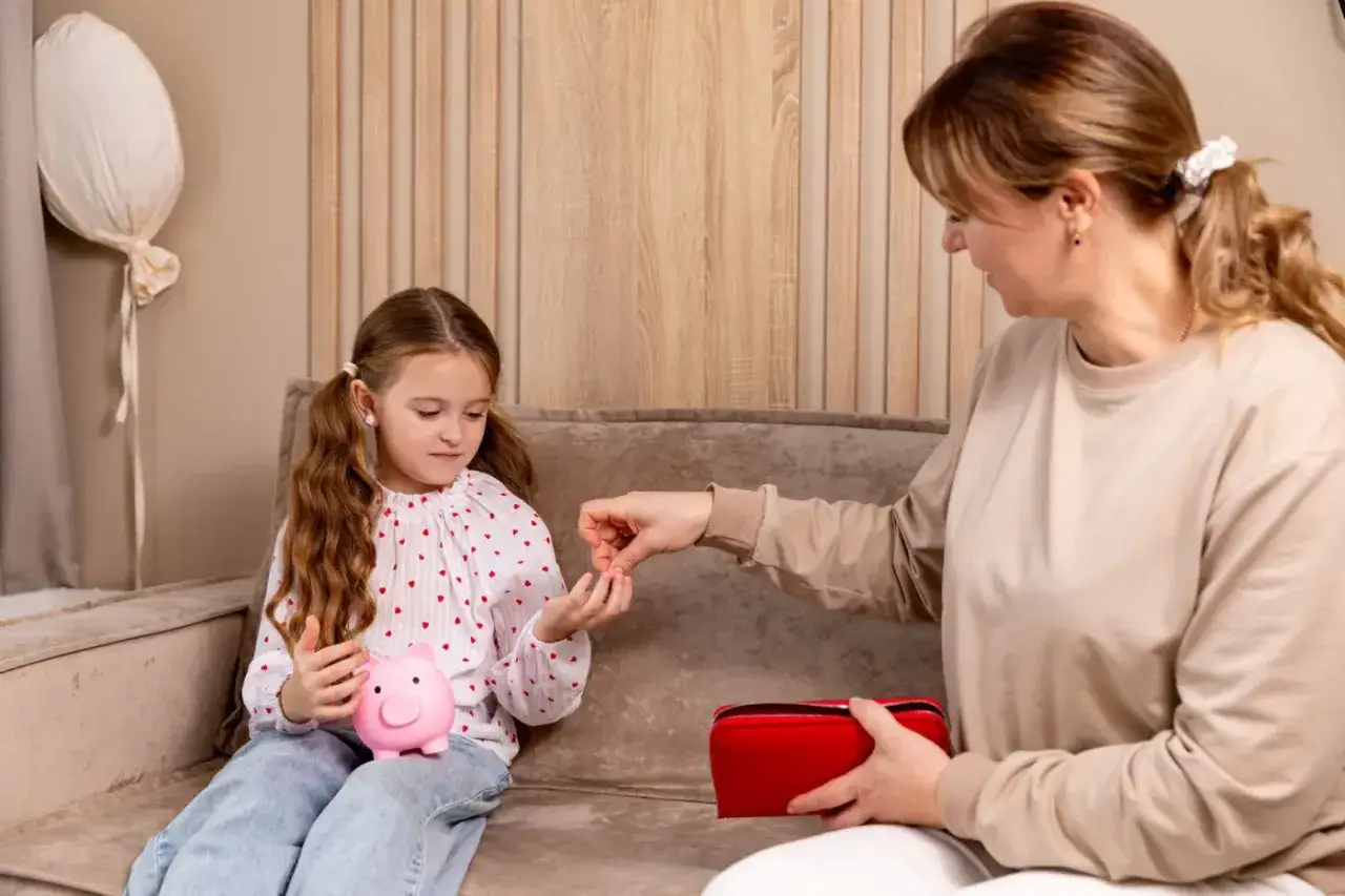Mama uczy córkę oszczędzać. Dziecko wkłada monetę do świnki-skarbonki. Zastanawiamy się, zasiłek macierzyński ile wynosi.