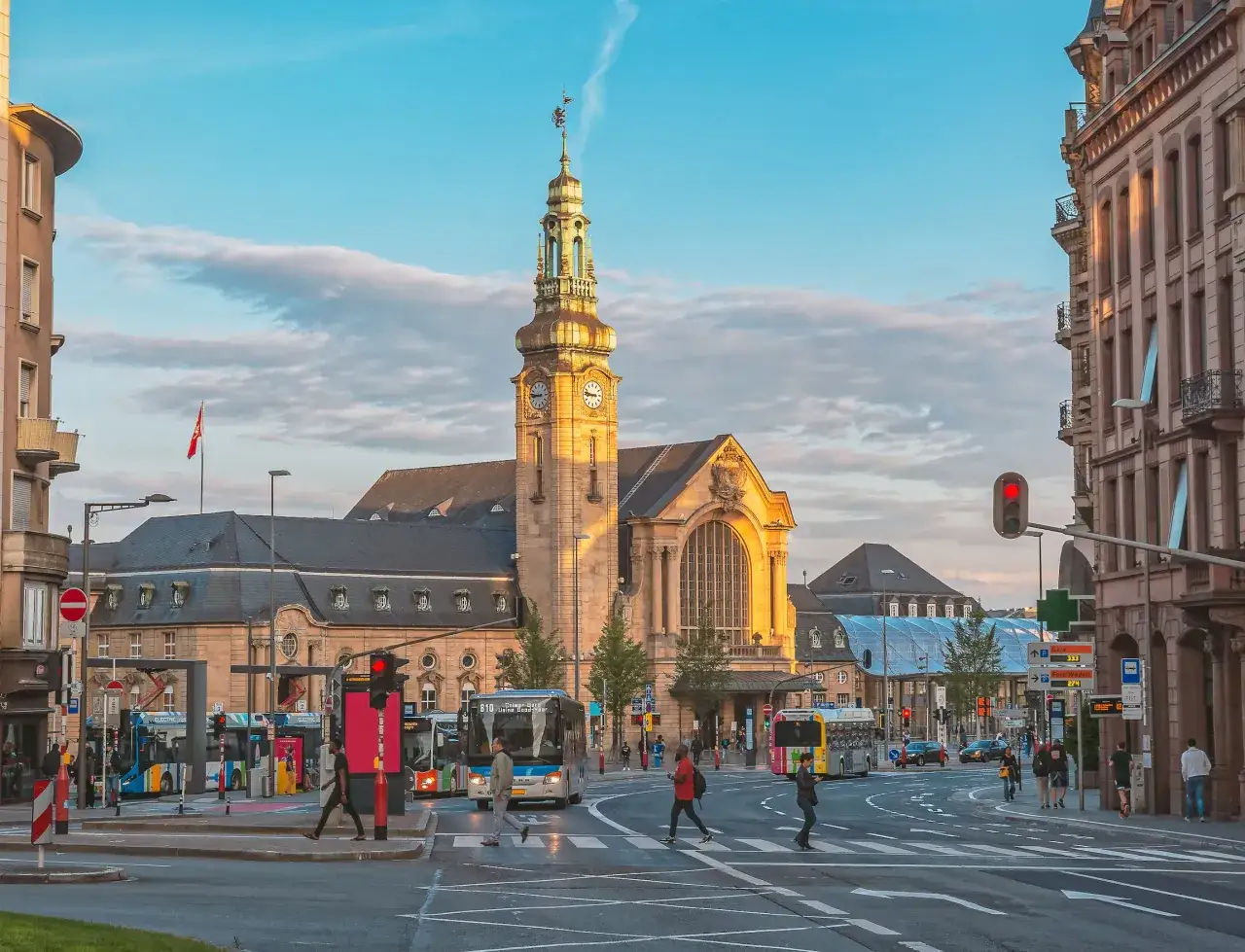 Ludzie spieszą się na autobusy w Luksemburgu, gdzie praca czeka na każdego. Złota wieża dworca góruje nad miastem.
