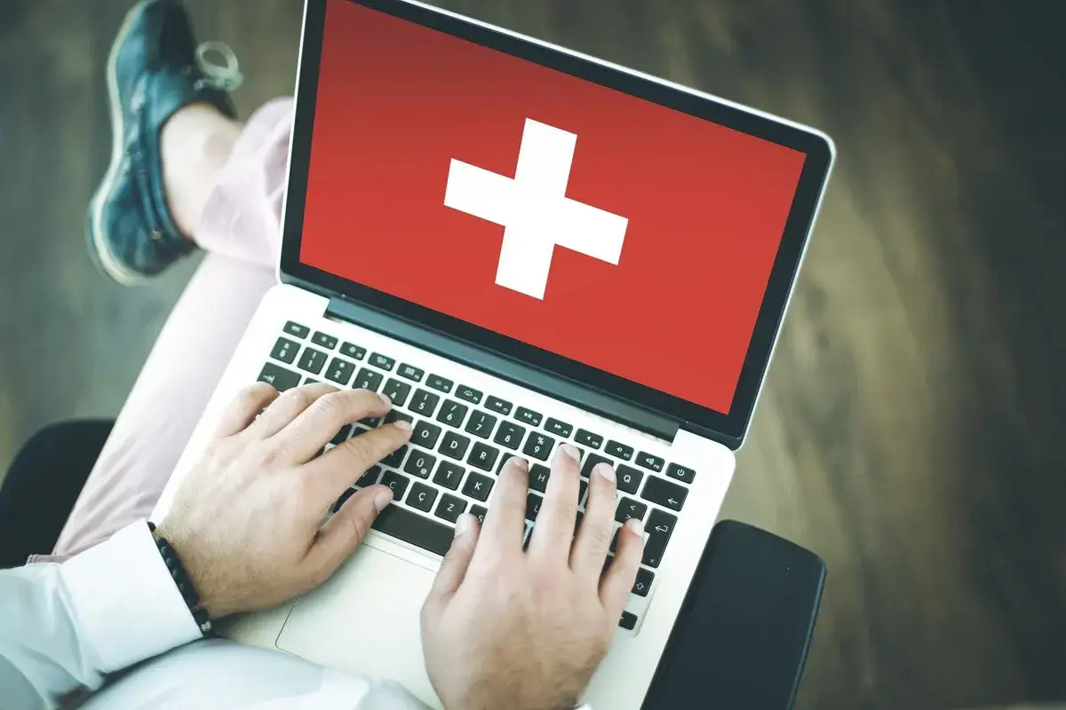 Osoba pracuje na laptopie z flagą Szwajcarii na ekranie. To może być początek nowej kariery lub poszukiwanie pracy w Szwajcarii.