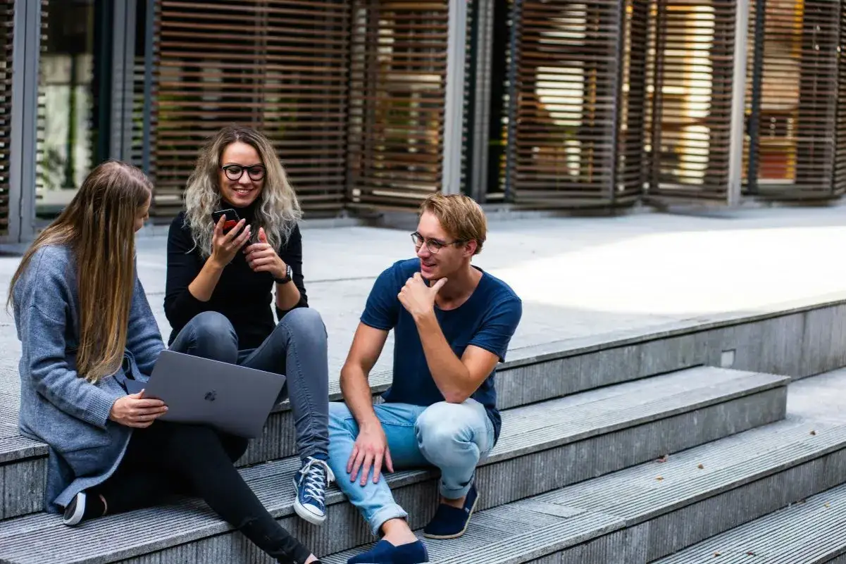 Trzech studentów, którzy wspólnie uczą się do egzaminów z języka angielskiego, siedzi na schodach. Jeden z nich trzyma laptopa, drugi telefon, a trzeci rozmawia z pozostałymi.