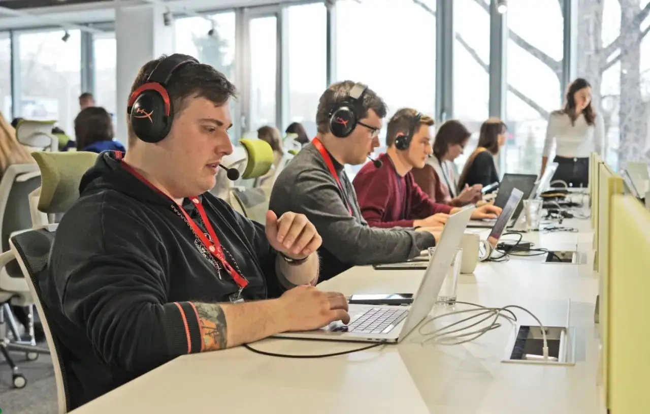 Młodzi ludzie w słuchawkach z mikrofonem pracują przy laptopach. To ich pmo stanowisko, gdzie skupienie jest kluczowe.