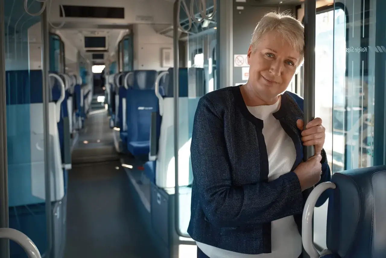 Uśmiechnięta kobieta w mundurze, gotowa do pracy jako konduktor w Kolejach Dolnośląskich. Wnętrze pociągu z niebieskimi siedzeniami.