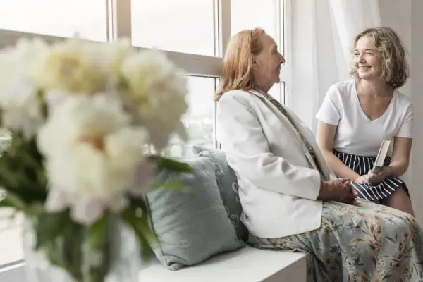 Uśmiechnięta młoda kobieta z książką rozmawia z seniorką przy oknie. Szukam pracy jako opiekunka prywatnie, oferując troskę i towarzystwo.