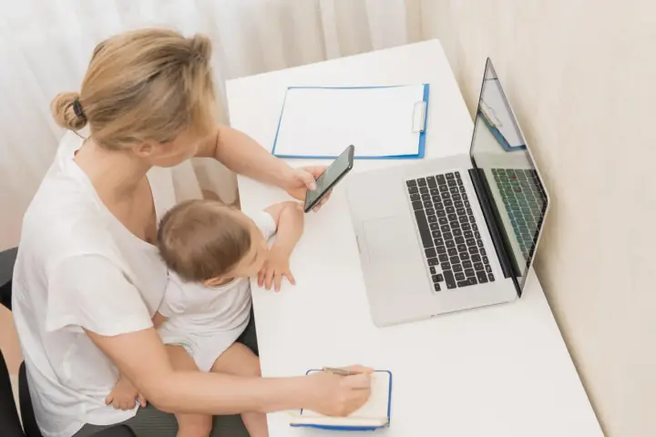Kobieta z dzieckiem na kolanach sprawdza na telefonie, zasiłek macierzyński ile wynosi, obok laptop i notatnik.