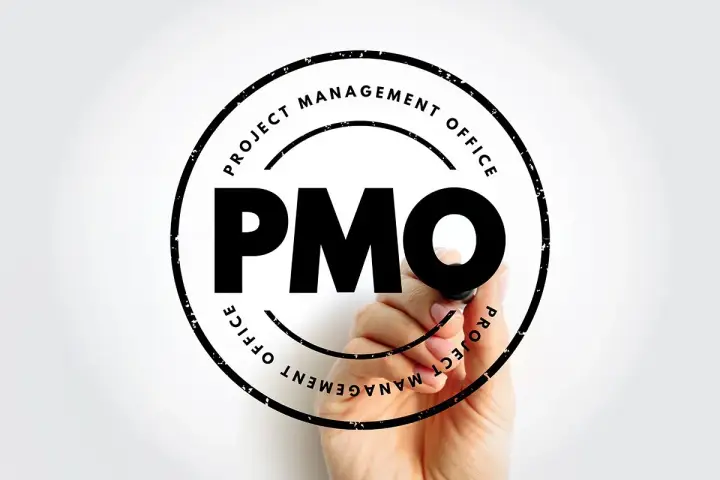 Ręka pisze markerem "PMO" w okręgu z napisem "PROJECT MANAGEMENT OFFICE". To symbol pmo stanowiska.