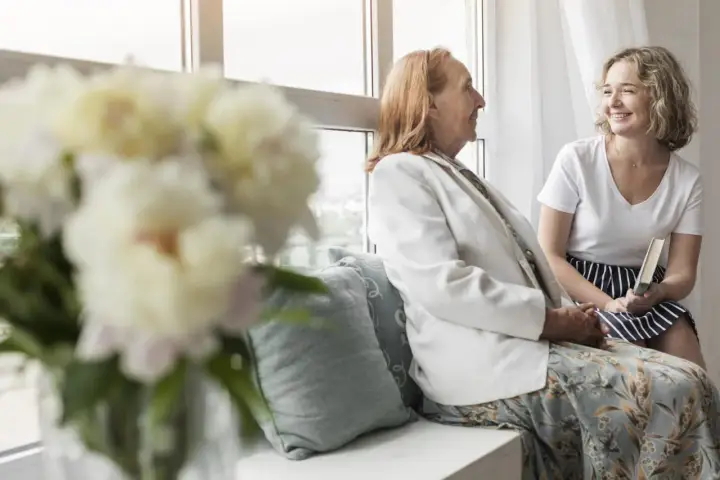 Uśmiechnięta młoda kobieta z książką rozmawia z seniorką przy oknie. Szukam pracy jako opiekunka prywatnie, oferując troskę i towarzystwo.